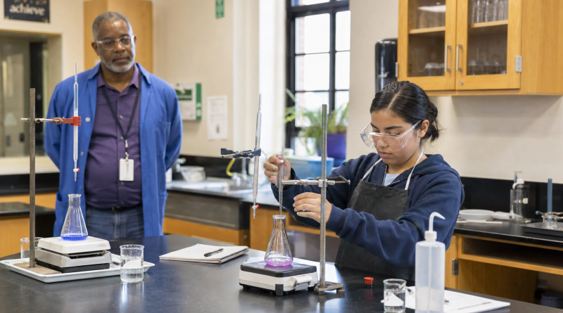 A student completes a science experiment independently while her teacher observes from several feet away