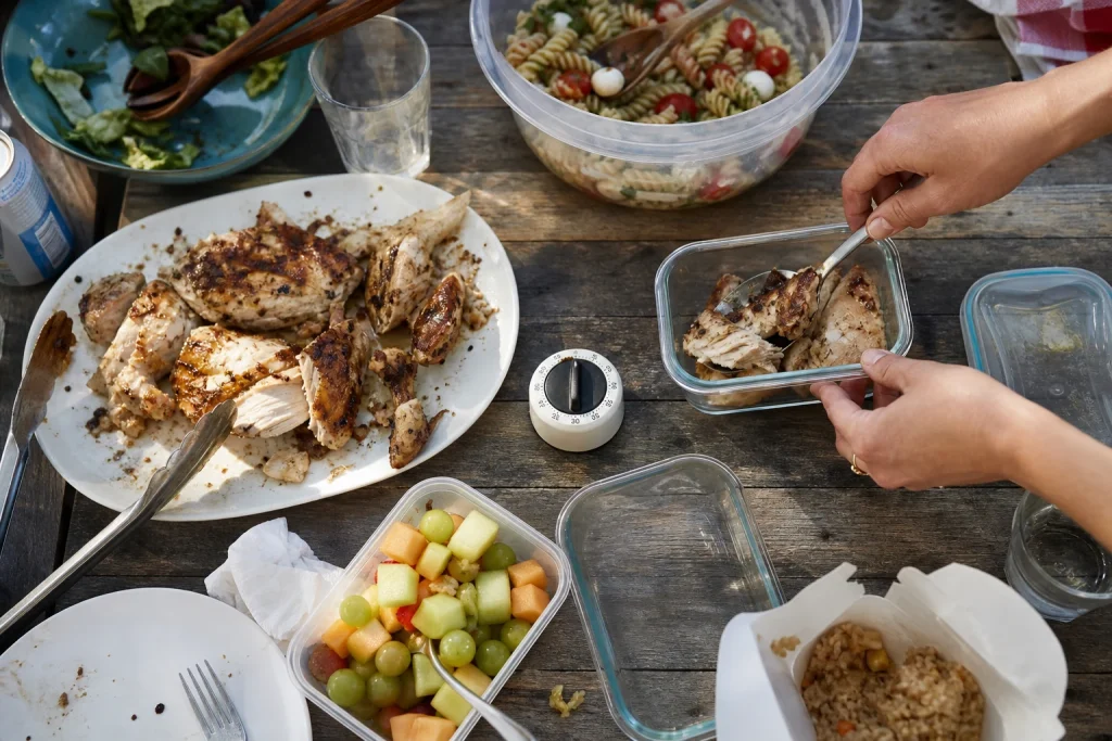 Hands pack cooked chicken and party food into shallow containers beside a kitchen timer