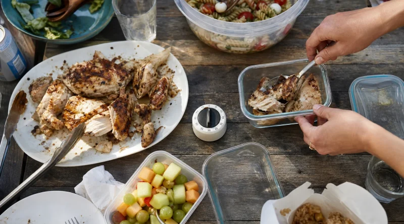 Hands pack cooked chicken and party food into shallow containers beside a kitchen timer